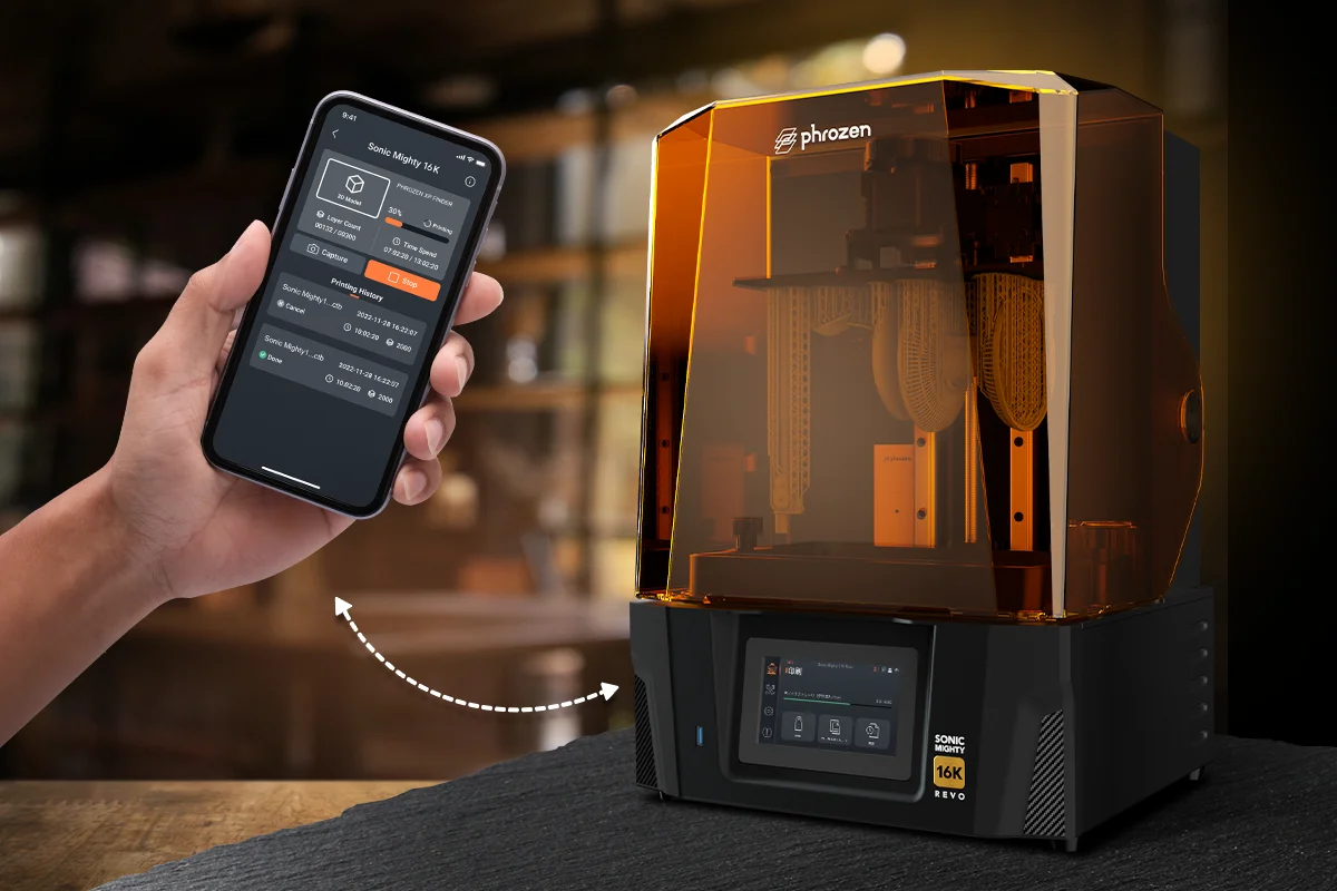 Person holds a smartphone displaying a 3D printer control app next to an orange-tinted Phrozen Sonic Mighty 16K 3D printer, implying remote control via mobile app.