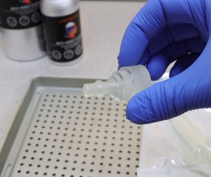 Gloved hand holding a clear plastic dropper nozzle over a perforated lab tray in a science setting.